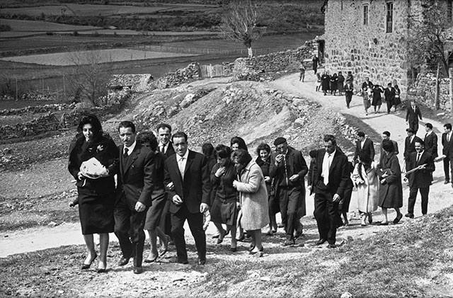 Una comitiva de boda asciende por un camino. Los primeros invitados quedan cerca de la cámara; otros todavía se agrupan junto al edificio del fondo.