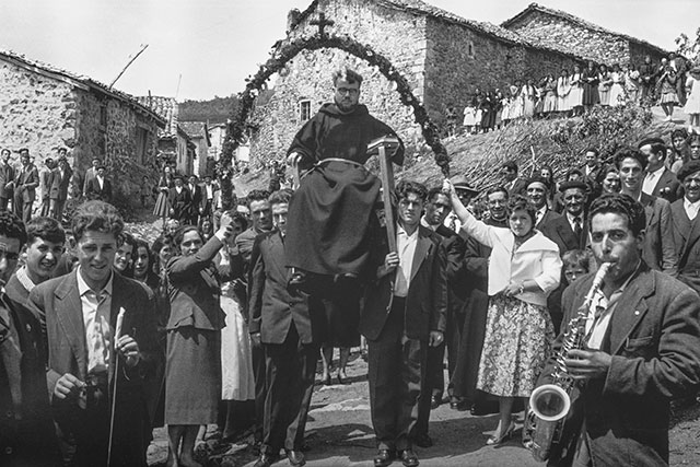 Un sacerdote es llevado bajo un arco adornado. Un músico con saxofón aparece en primer plano; el público ocupa la calle y el desnivel del fondo.