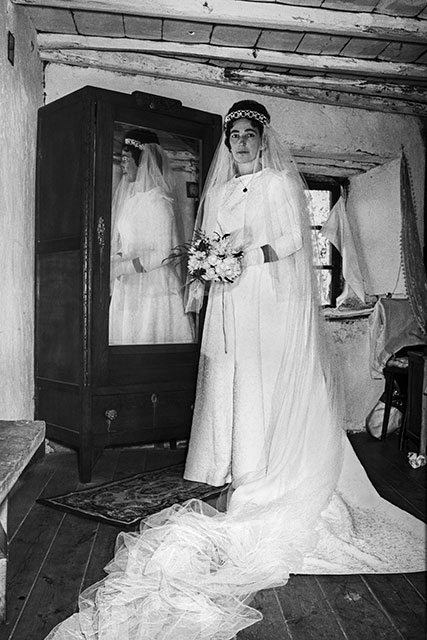 Emilia posa con vestido y velo. El espejo de un armario recoge otra vista de ella; la cola del vestido ocupa el suelo de la habitación.