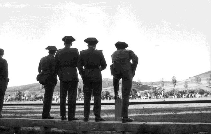 Varios guardias civiles, vistos de espaldas, observan un espacio abierto. Sus siluetas oscuras se recortan contra el cielo y el fondo lejano.