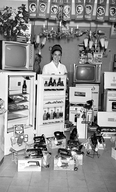 Una mujer sonríe entre televisores, frigoríficos abiertos, planchas y lámparas. Los objetos llenan el escaparate interior de la imagen.